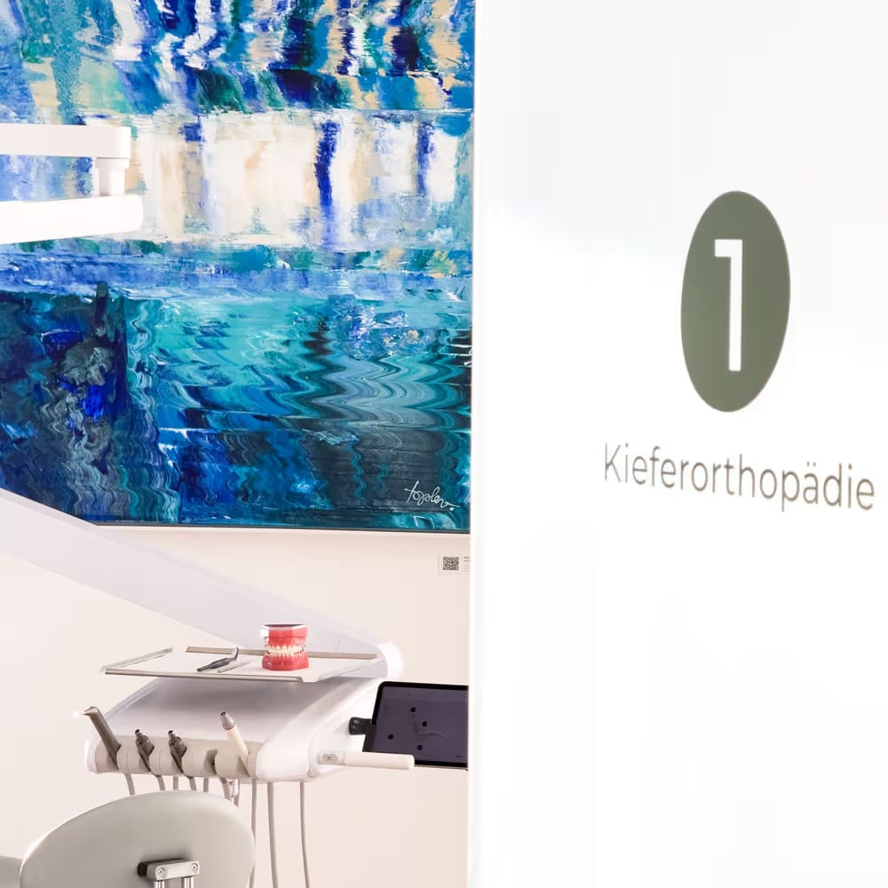 Zahnmedizinischer Behandlungsstuhl und Gerät vor abstraktem blauen Kunstwerk in Praxis mit Türschild Nummer 1 und Aufschrift Kieferorthopädie.