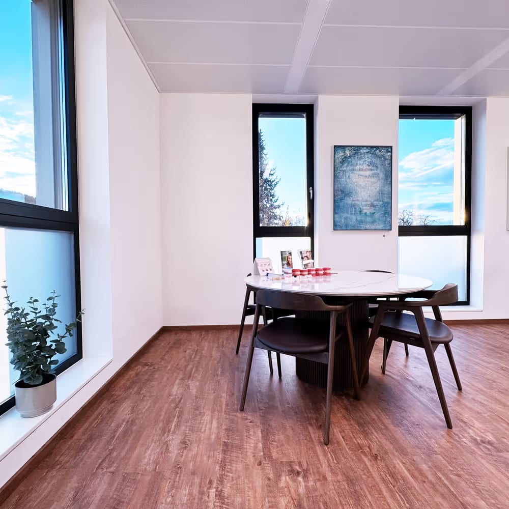 Moderner Besprechungsraum mit rundem Tisch, vier Stühlen, großen Fenstern und einem kleinen Topf mit Pflanze am Fenster.