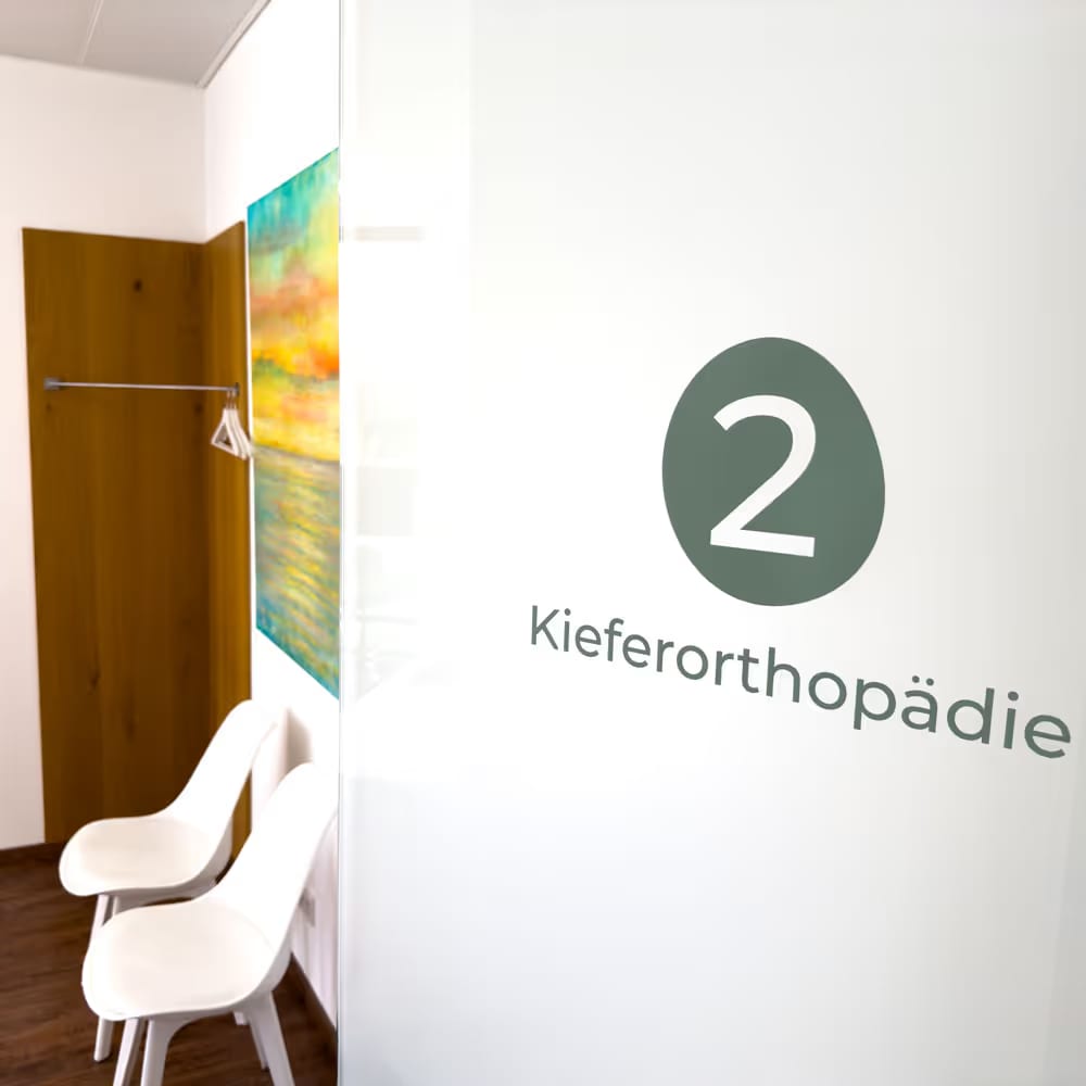 Behandlungsraum mit zwei weißen Stühlen und einem bunten Gemälde an der Wand dahinter.